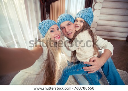 holidays, technology and people concept - happy family sitting on floor and taking selfie picture with smartphone at home, in a winter sweater and hat, the concept of Christmas