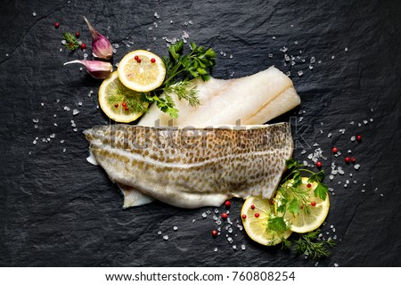 Fresh fish,  raw cod fillets with addition of herbs and lemon slices on black stone background, top view                       