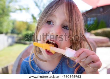 portrait close up of face of four years old blonde girl eating orange or yellow ice lolly or popsicle