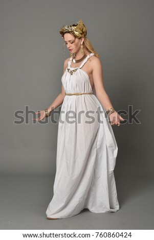 full length portrait of girl wearing white ancient Greek or Roman costume, standing pose on a grey background.
