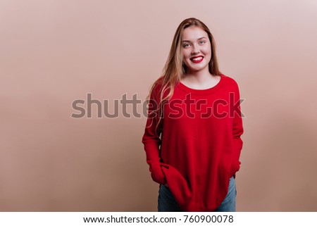 Gorgeous long-haired girl with red lips wearing red pullover posing on beige background with lovely charming smile. Portrait of friendly woman with truly happy emotions, place for text