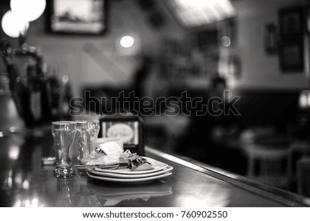 Focused set of used cutlery after breakfast in a cafe bar.Black and white indoor picture with blurred background and glasses, plates and other stuff at the foreground.Sad, hurry and loneliness concept