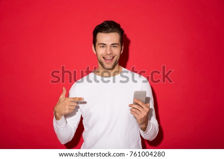 Picture of smiling man in sweater looking at the camera while holding smartphone and pointing at him over red background
