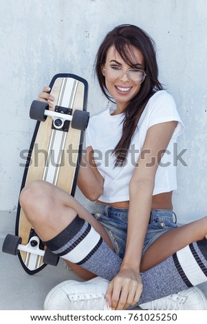 Happy young fashionable trendy woman sitting with skateboard at wall