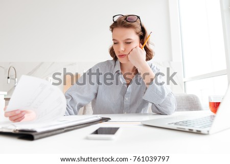 Attractive thinking woman in striped shirt holding her cheek while siting and reading contract at home
