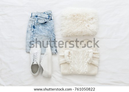 Blue jeans, white knitted sweater, sneakers, fur scarf  lying on bed on white sheet. Overhead view of woman's casual outfit. Trendy hipster look. Women winter or spring clothes. Flat lay, top view.