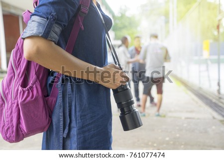 Close up woman photographer hand, wearing blue jens dress and pink back pack, holding camera with long zoom lens with tennis court background. Travel and hobby concept.
