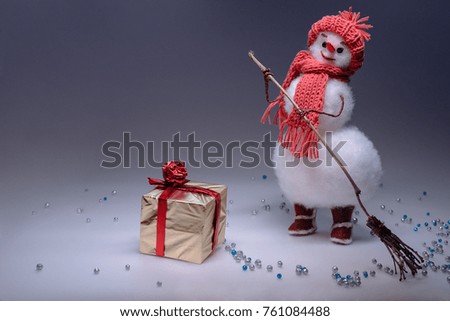 A snowman in a hat and scarf sweeps the snow with a broom and looks at the box with a gift. Smooth gradient background. Snowfall.