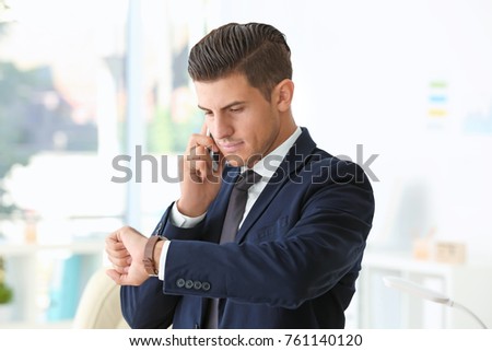 Handsome businessman talking on mobile phone and looking at watch in office