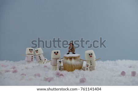 Christmas food photography picture using marshmallows shaped as cute snowman with iced on smiles and standing in snow with cream fairy cake and Santa Claus decoration on top 
