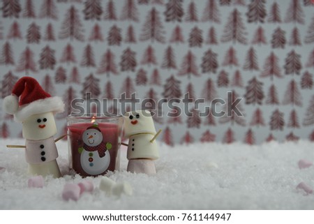 Christmas photography image of red  candle with lit flame and marshmallows shaped as snowman with iced on smiles standing in snow with shiny red tree pattern background