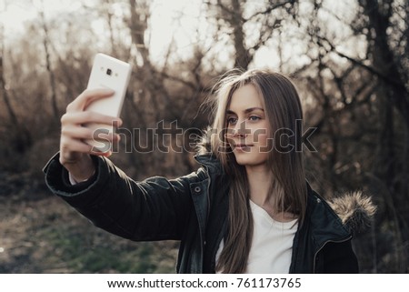 Pretty young woman taking selfie on mobile outdoor. Beautiful girl about twenty eyers old posing at camera in park. Model dressed in parka. Woman with blonde hair, full lips, wide eyebrows, blue eyes.