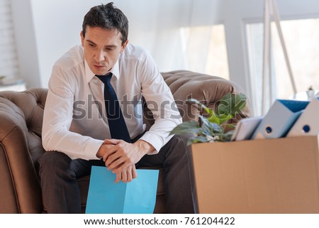 Thinking over. Clever concentrated serious man sitting in an armchair with an important document in his hand and thinking about his lost job while a big box with personal items standing by his side