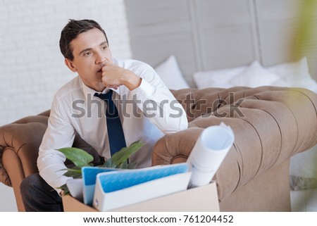 Difficult decision. Clever experienced specialist sitting in an armchair with a box of personal items by his side and looking into the distance while thinking of leaving his work