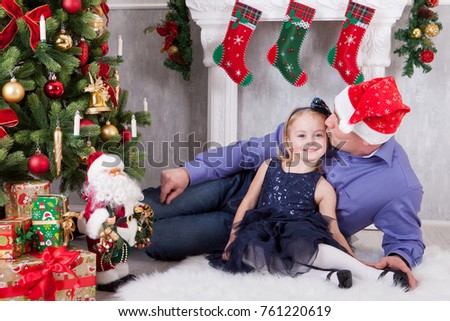 Christmas or New year celebration. Father kisses the daughter near the Christmas tree. Father have a Santa Claus hat on his head. A fireplace with christmas stocking behind them