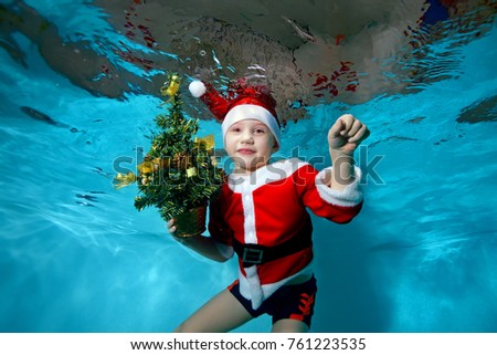 Happy child in Santa Claus hat swims and poses underwater in the pool with Christmas tree in hand, looking at camera and smiling. Portrait. Shooting under water. Horizontal orientation
