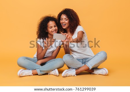 Portrait of a two joyful afro american sisters taking a selfie while sitting and showing peace gesture isolated over orange background