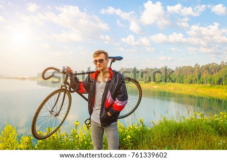 man stand with bicycle in hands on the hill and looking at the lake with yellow flowers and beautiful sunrise