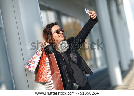 Woman in sun glasses a black leather jacket, black jeans Holding a Bunch of Shopping Bags Doing Selfie in front of mirrored windows on the Street