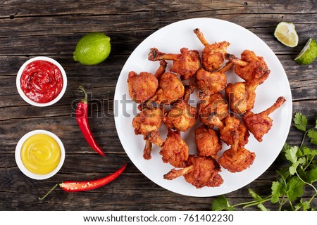 delicious deep fried battered crispy chicken winglets  with exposed bones on a white plate on dark wooden table with mustard and tomato sauce in gravy boats at background,, horizontal view from above Royalty-Free Stock Photo #761402230