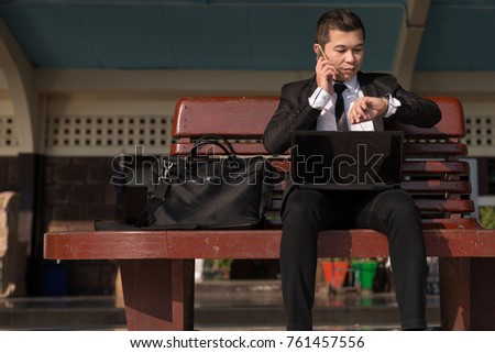 Businessman sitting at train station is looking at watch in hand holding mobile phone. Networking Partnership Communication Business