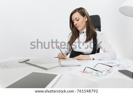Beautiful serious brown-hair business woman in suit and glasses sitting at the desk, working at computer with modern monitor and documents in light office, writing in notebook on white background.
