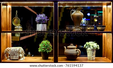 different beautiful artificial flowers in pots, ceramic jar with mugs, teapot, elephant statue on a wooden shelf decorated with lights