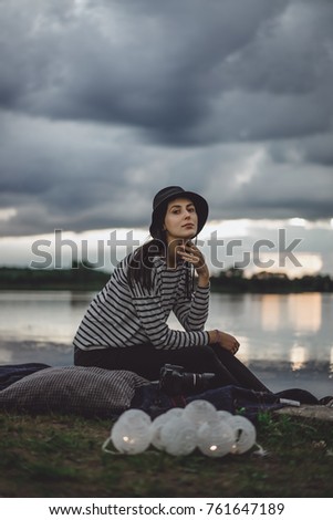beautiful brunette girl in hat and striped blouse at sunset by the water on the lake shore.