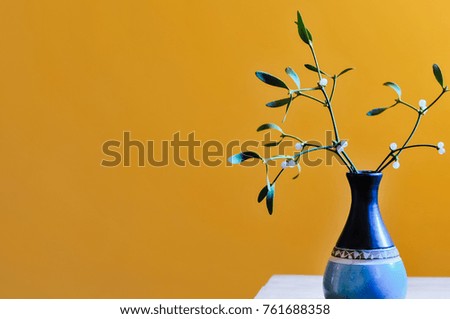 Mistletoe twig with leaves and white berries in blue, hand pottered vase on wooden table in front of orange-yellow background with lots of copy space