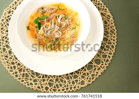 Venison soup with noodle, carrot and parsley in white plate, circle bamboo placemat on green background. 