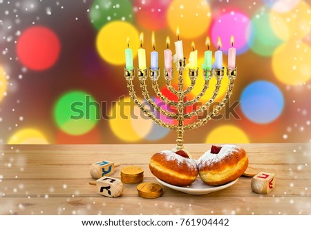 Menorah with candles, donuts, golden chocolate coins and wooden dreidels with red, blue, black letters on wooden table on blurred bokeh background of colored lights. Jewish holiday Hanukkah.