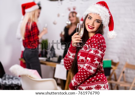 Portrait of a young woman with a glass of champagne at home on the foreground. Beautiful brunette in a Santa hat, red costume with deers