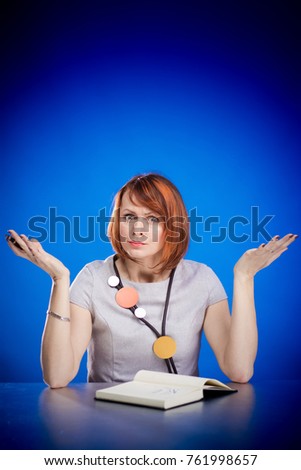 Emotional red-haired businesswoman sitting at table and posing on blue background
