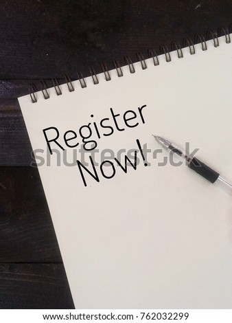 The letter REGISTER NOW! on notebook with pen. Wood background