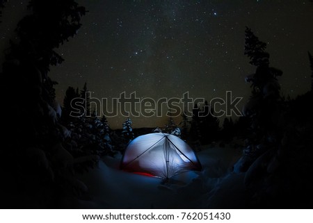 White tent under the starry sky and the milky way in the trees in the winter mountains at night.