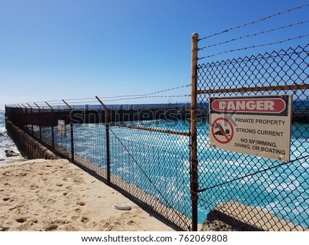 Sectioned off pretty blue beach surrounded by sand and a barbed fence, displaying a sign warning of the dangers of swimming there. 