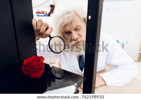 An elderly man is a scientist in a laboratory room. The man is experimenting with a 3D printer. A scientist with a gray beard looks through a magnifying glass on a rose in a 3D printer.