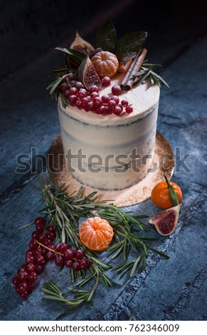 Angle shot of white christmas cake decorated with red berries, tangerin, figs, cinnamon stick and rosemary