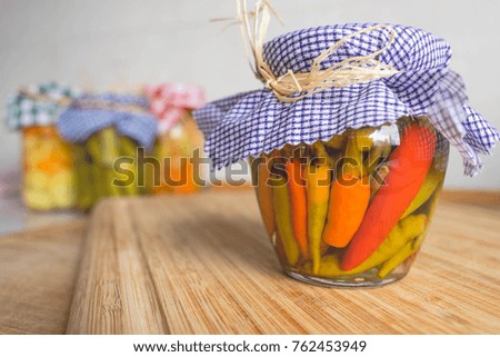 A small jar of pickled vegetables with a blue and white checkered fabric top on wooden background and blurred kitchen background, vintage toned