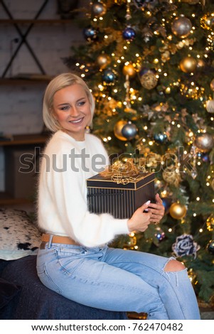 Beautiful happy girl with short blonde hair is holding a New Year gift/ girl is dressed in a fluffy white sweater and blue jeans/beautiful Christmas tree on background