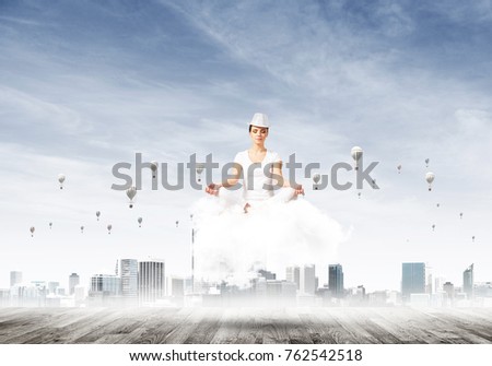 Woman in white clothing keeping eyes closed and looking concentrated while meditating on cloud with cityscape view and flying balloons on background.