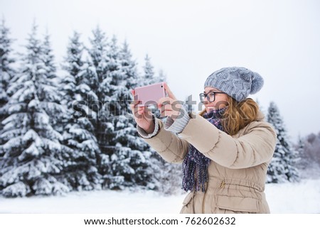 Young woman takes photo on a background of snow-covered winter forest. Snowy weather. Woman taking  photo on phone. Girl smiling at the camera. Winter holidays.