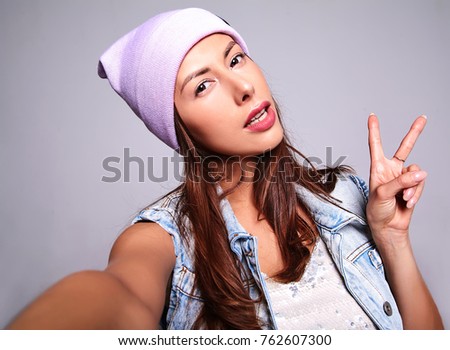 Portrait of beautiful cute brunette woman model in casual summer jeans clothes with no makeup in purple beanie making selfie photo on smartphone isolated on gray. Showing peace sign
