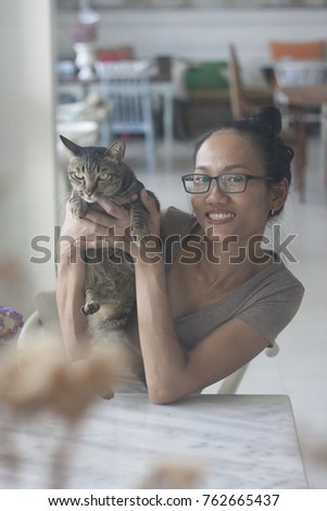 Asian Lady Women Girl holding hugging cat with a big smile sitting on white wishbone chair in home cafe behind white marble table. Picture was taken through glass window