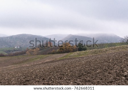 Autumn rural landscape with plowed agricultural field and vineyard in the foreground and house and foggy mountains in the background on a cloudy autumn afternoon