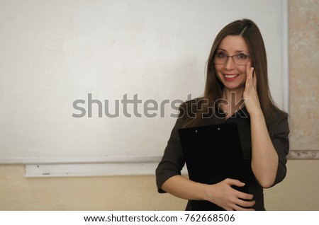 Young teacher in glasses with day book stands and smiles in a school class on school board background with copy space. Education concept. 