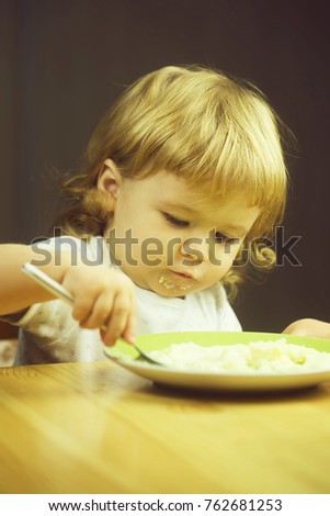 Closeup portrait view of one adorable cute small baby boy with blonde hair eating healthy food of porridge or coocked semolina from plate with spoon in hand indoor, vertical picture