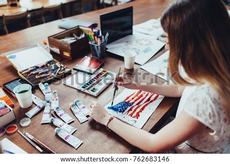 Young female illustrator drawing flag of the USA in using aquarelle paint sitting at workplace