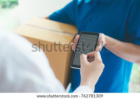 Young woman appending signature in digital mobile phone after receiving parcel from courier at home. Royalty-Free Stock Photo #762781309