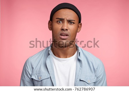 Portrait of discontent mixed race young male wears black hat and shirt, frowns face, looks puzzled, isolated over pink background. Hipster guy in bewilderment. People and facial expressions concept